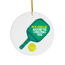 Load image into Gallery viewer, The Pickle Ball Christmas Christmas Ornament by R is for Robo features a white ceramic design adorned with a green pickleball paddle and the festive text "PICKLEBALLIN AROUND THE CHRISTMAS TREE" in yellow and white. Beneath the paddle rests a yellow pickleball, and it includes a gold string for easy hanging, making it the ideal pickle ball holiday ornament.