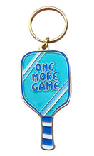Load image into Gallery viewer, The Pickleball Paddle Enamel Keychain, by R is for Robo, is designed to resemble a blue pickleball paddle with the phrase "ONE MORE GAME" prominently displayed in white. The handle showcases various shades of blue stripes and is paired with a gold-colored key ring for convenient attachment.
