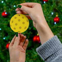 Load image into Gallery viewer, With their green-painted nails, a person hangs the Pickleball Ball Christmas Ornament - Ceramic Holiday Ornament by R is for Robo on a Christmas tree decorated with red baubles. The person, dressed in a gray sweater, stands against a blurred festive background.