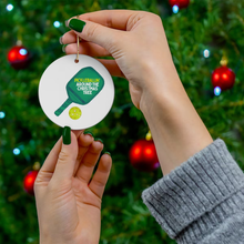 Load image into Gallery viewer, A person with green-painted nails holds the "Pickle Ball Christmas Christmas Ornament" by R is for Robo, a ceramic holiday decoration showcasing a pickleball paddle and ball illustration, along with the phrase "Pickleballin Around the Christmas Tree." The ornament shines brilliantly in front of a tree festooned with red baubles, perfectly capturing the holiday spirit.