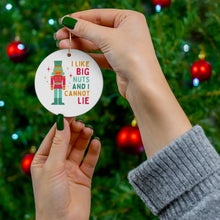 Load image into Gallery viewer, A person with green nail polish holds a humorous Christmas ornament from R is for Robo's collection, featuring a nutcracker and the text "I like big nuts and I cannot lie." In the background, a Christmas tree is adorned with red baubles. This festive decoration adds a playful touch to your holiday decor.