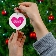 Load image into Gallery viewer, Wearing green nail polish, a person holds R is for Robo's Cunt Christmas Ornament - Funny Gifts for Women, featuring a striking pink heart with bold text. The ornament stands out against a festive backdrop of a decorated Christmas tree adorned with red baubles.