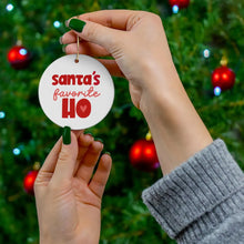 Load image into Gallery viewer, In front of a lit Christmas tree, two hands hold a playful holiday gift—R is for Robo's Santa's Favorite Ho - Funny Christmas Ornament for Her, featuring decorative red text and a red heart symbol. This ornament brings a sassy flair to the holiday décor.