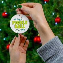 Load image into Gallery viewer, In a festive scene, two hands present the "In my Pickleball Era Pickleball Christmas Ornament" by R is for Robo in front of a decorated tree. The ornament displays the phrase in green and yellow letters. The tree is adorned with red baubles, while the person wears a gray sweater and matching green nail polish, enhancing the holiday atmosphere.