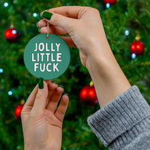 Load image into Gallery viewer, A person with green nail polish holds the "Jolly Little Fuck Funny Christmas Ornaments for Him or Her" by R is for Robo, featuring cheeky white lettering. The backdrop displays a festive Christmas tree embellished with red and blue decorations, making it a sassy holiday gift idea for fans of irreverent seasonal decor.