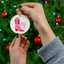 Load image into Gallery viewer, A person with green nail polish holds a round "Pink Cowboy Boot Christmas Ornament for Women" by R is for Robo, which showcases a pink cowboy boot and the phrase "Merry Christmas y'all." In the background, a Christmas tree adorned with vibrant red baubles enhances the festive scene.