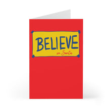 Load image into Gallery viewer, The Ted Lasso Believe in Santa Christmas Card by R is for Robo showcases a red background with a yellow rectangle and a blue border. Inside, the word BELIEVE is prominently displayed in large blue letters, with "in Santa" added below in smaller red script, exuding holiday cheer as the card stands open.