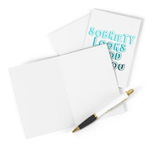 Load image into Gallery viewer, A blank turquoise card and an unopened Sobriety Soberversary Congratulations Card by R is for Robo with "Sobriety Looks Good on You" in teal letters are displayed. A black and gold pen rests on top of the open card.