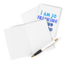 Load image into Gallery viewer, A blank greeting card from R is for Robo, accompanied by a black and gold pen, rests beside a heavyweight matte card titled "I am So Freaking Proud Congratulations Card." The card showcases the message "I AM SO FREAKING PROUD OF YOU" in vibrant blue letters, making it ideal for personalizing with your own congratulatory message.