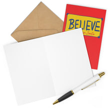 Load image into Gallery viewer, A Ted Lasso Believe in Santa Christmas Card from R is for Robo lies open alongside a brown envelope, embodying the spirit of holiday joy. Its red design is emblazoned with the "BELIEVE" message in yellow and blue, echoing Ted Lasso's cheerful optimism. A white pen with a gold accent rests diagonally across the card.
