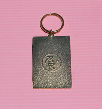 Load image into Gallery viewer, The Cassette Tape Keychain - Mix Tape Keychain for 90s Kid by R is for Robo captures a retro flair with its metallic rectangular design featuring a round emblem of abstract symbols. Attached to a simple gold keyring and placed on a textured pink surface, it offers a touch of vintage charm.