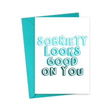 Load image into Gallery viewer, Featuring a teal envelope, the Sobriety Soberversary Congratulations Card by R is for Robo proudly displays "Sobriety Looks Good On You" in bold, playful turquoise and dark blue letters on a crisp white background.
