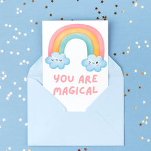 Load image into Gallery viewer, A delightful thank you card from R is for Robo, featuring a rainbow and smiling clouds design, peeks out of a light blue envelope. The text "You Are Magical" is printed in pink beneath the rainbow on a blue background adorned with scattered gold star confetti.