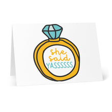 Load image into Gallery viewer, The She Said Yas Funny Engagement Card by R is for Robo features a delightful design of a prominent gold ring with a blue diamond. Inside, whimsical yellow and teal letters excitedly announce "she said YASSSSSS." The card stands upright on a white surface, making it ideal for adding your own personal message.