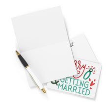 Load image into Gallery viewer, The Merry & Getting Married Engagement Christmas Card by R is for Robo features a blank card and envelope beside a pen, waiting to be personalized. The smaller card reads 'Getting Married' in festive green and red, with a charming ring and heart design that adds a delightful twist reminiscent of a Christmas card.