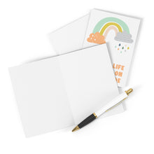 Load image into Gallery viewer, A blank sympathy card with a gold and black pen lies next to the "Rain on Your Parade - Cute Sympathy Card" from R is for Robo, which is a vibrant card adorned with a rainbow, clouds, and raindrops. The text on the card reads Life on in pastel colors.
