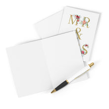 Load image into Gallery viewer, The elegant Mr and Mrs Wedding Card from R is for Robo, featuring gold lettering and a floral design, lies open, inviting a handwritten note. A nearby black and gold pen waits patiently to capture personal thoughts.