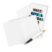 Load image into Gallery viewer, A partially open white birthday card rests on a white surface accompanied by a black and gold pen. In the background, there's the Stoner Birthday Card Wake Bake & Birthday Cake from R is for Robo, featuring vibrant text that reads, "Wake, Bake, & Birthday Cake," making it an ideal stoner gift for those who enjoy humor.